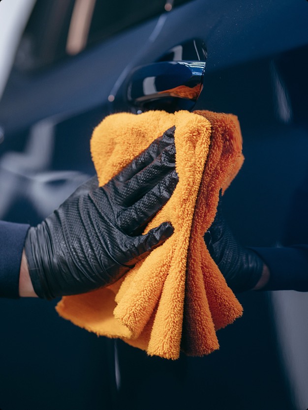 Mano con guanto nero che tiene un panno arancione, mentre pulisce la superficie di un'auto. L'immagine mostra attenzione ai dettagli e cura nella pulizia.