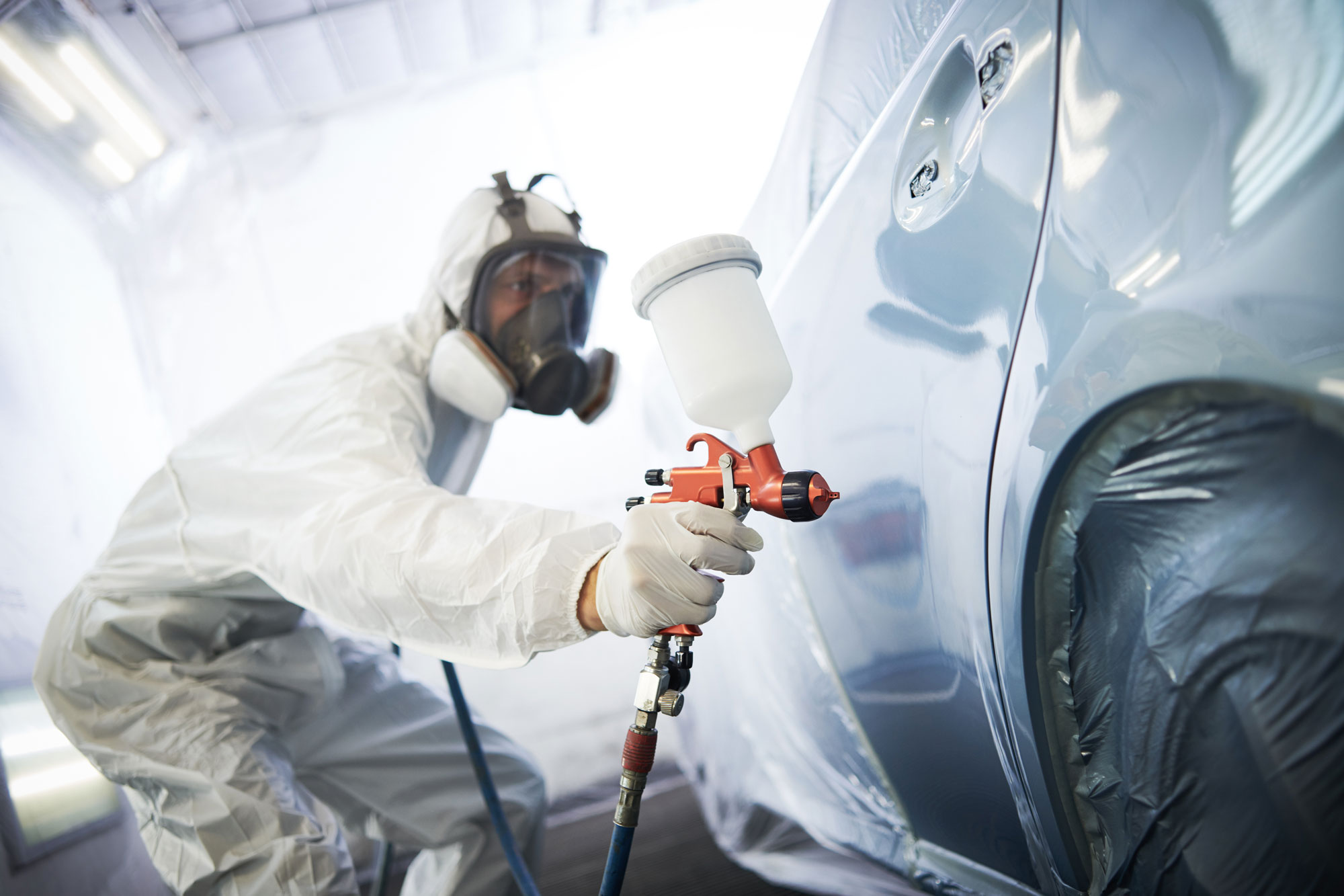 Un professionista in tuta protettiva e maschera spruzza vernice su un'auto blu, all'interno di un garage specializzato per verniciatura.