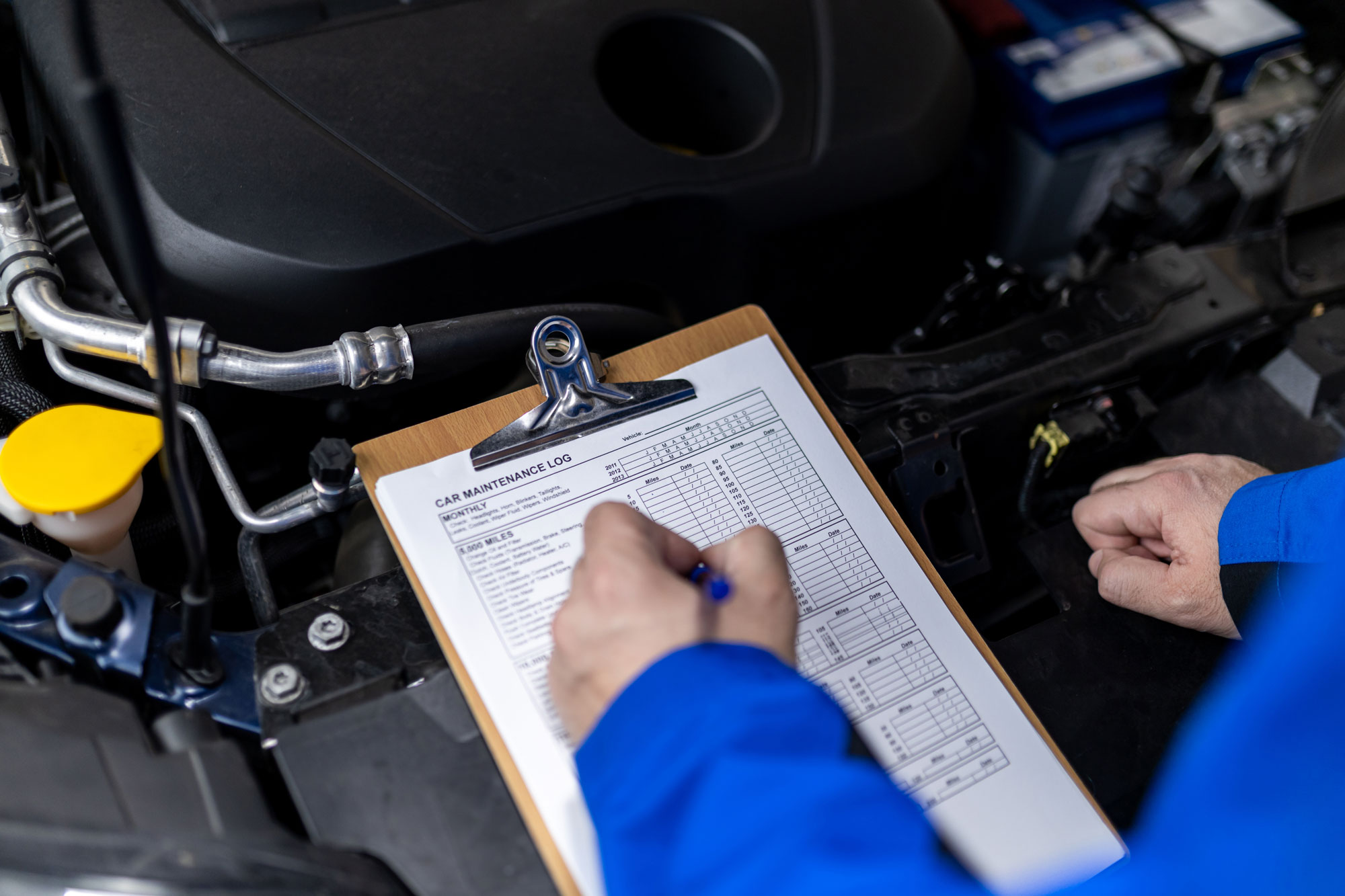Un meccanico sta prendendo appunti su una clipboard mentre controlla il motore di un'auto. Il foglio contiene una lista di controllo per la manutenzione.