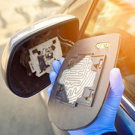 Un tecnico tiene in mano un componente di specchio retrovisore automobilistico, mentre lavora su un veicolo. Il pezzo mostra dettagli intricati. La luce naturale illumina l'immagine.